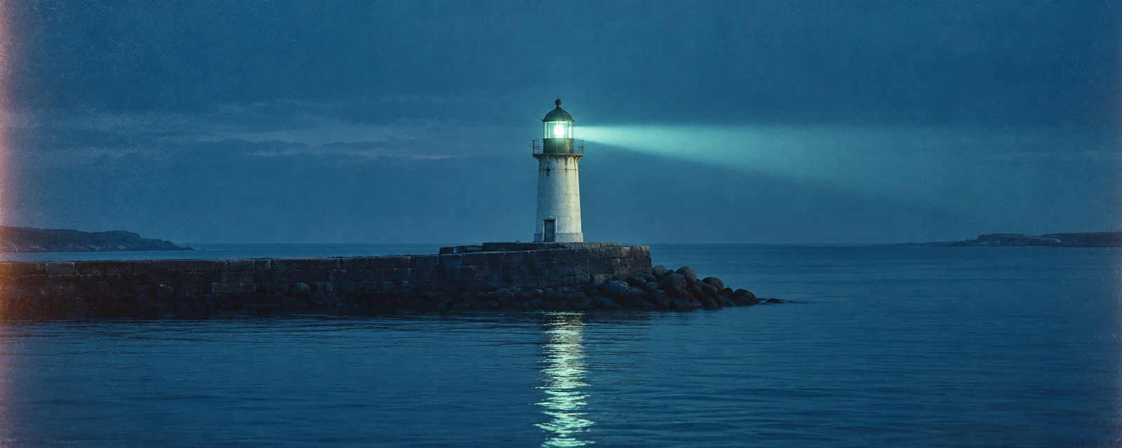 Vintage film photograph: A lighthouse on a stone breakwater casts a beam across the sea in blue twilight.